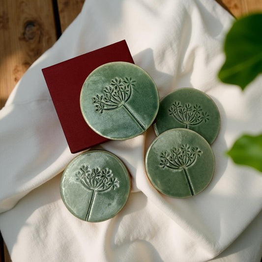 A set of four green ceramic coasters with a floral pattern, displayed on a white fabric surface with a red box in the background.